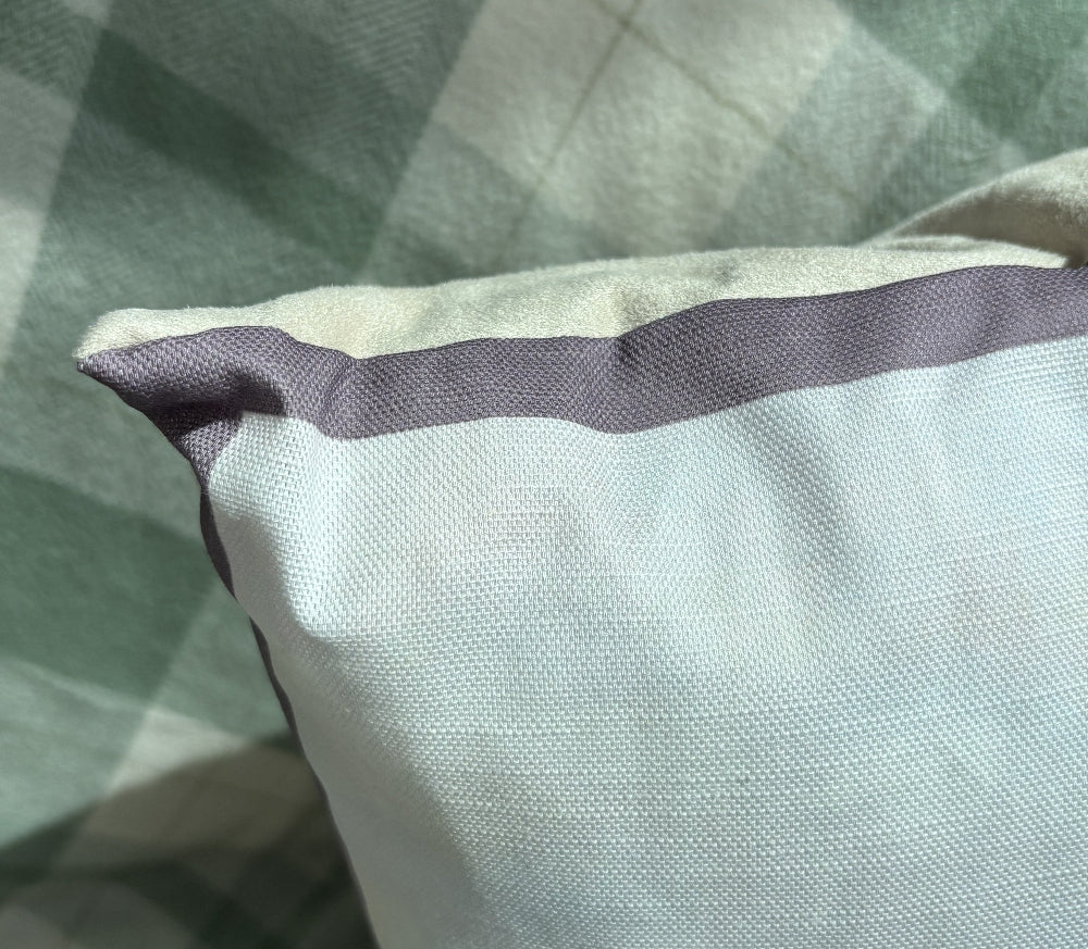 A close-up of a light gray fabric pillow or cushion with a contrasting dark gray horizontal stripe and a pointed corner, resting on a green and white plaid blanket.