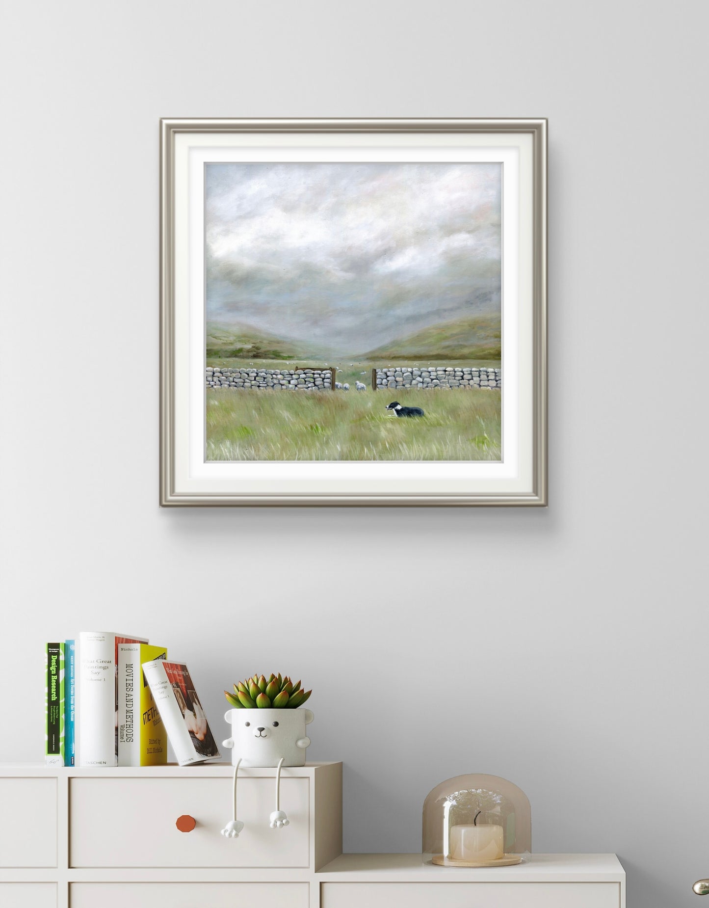 Framed landscape painting on a wall above a white cabinet with decorative items.