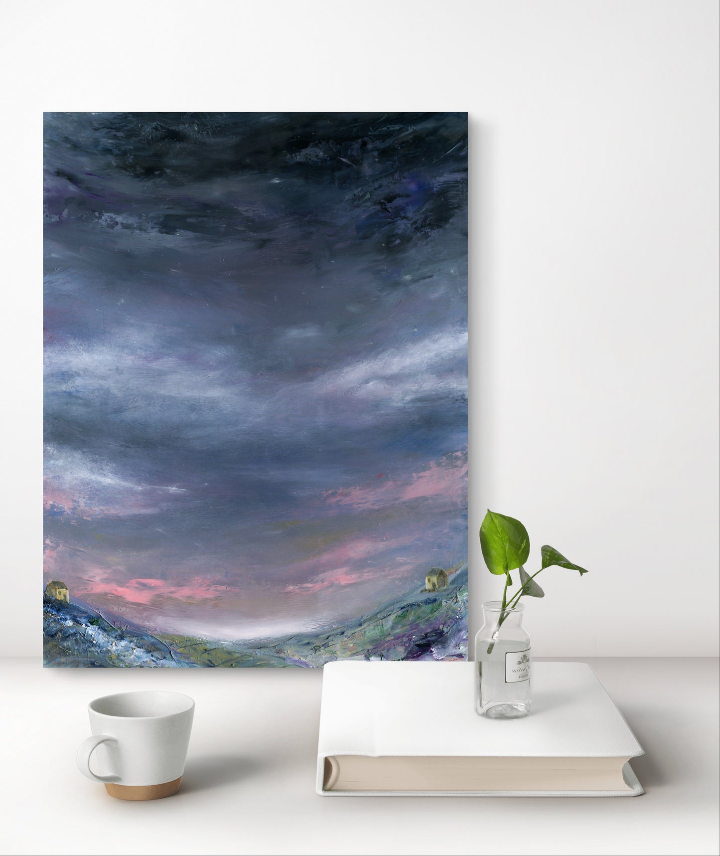 Art print of a landscape with a purple sky on a white surface with a cup, book, and plant.