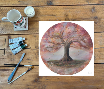 A circular art print featuring a painted tree with pink, red, and brown autumn leaves, displayed on a rustic wooden table alongside painting supplies including a mug, candle, paint tubes, brushes, and a palette.