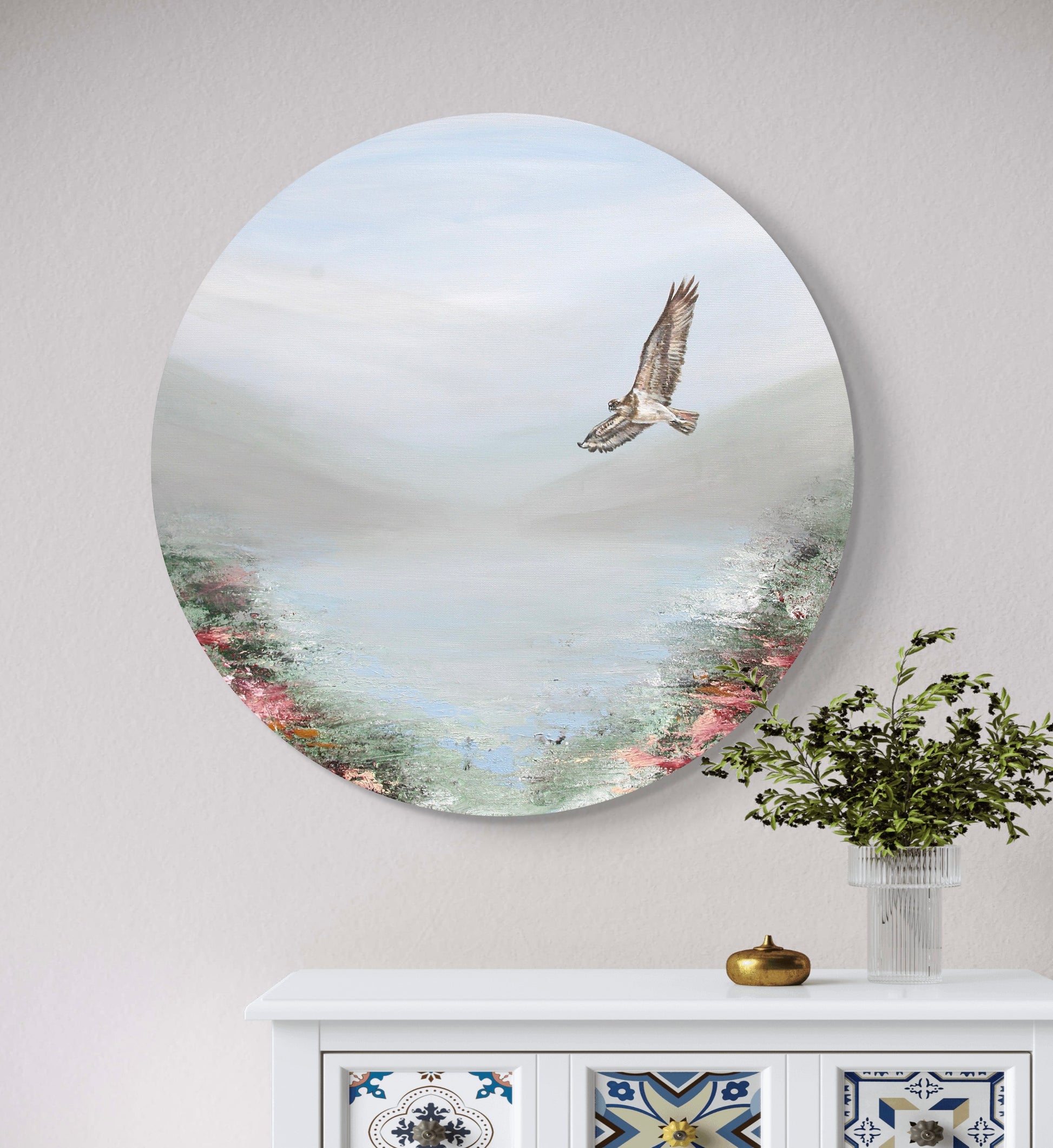 An acrylic painting depicting an osprey flying over a loch, with a serene, misty background. The painting is round and has a frame, mounted on a wall above a white cabinet with a vase of flowers.