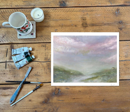 A landscape art print with a white border is displayed on a rustic wooden table alongside painting supplies, including a floral mug, a candle, paint tubes, and brushes.