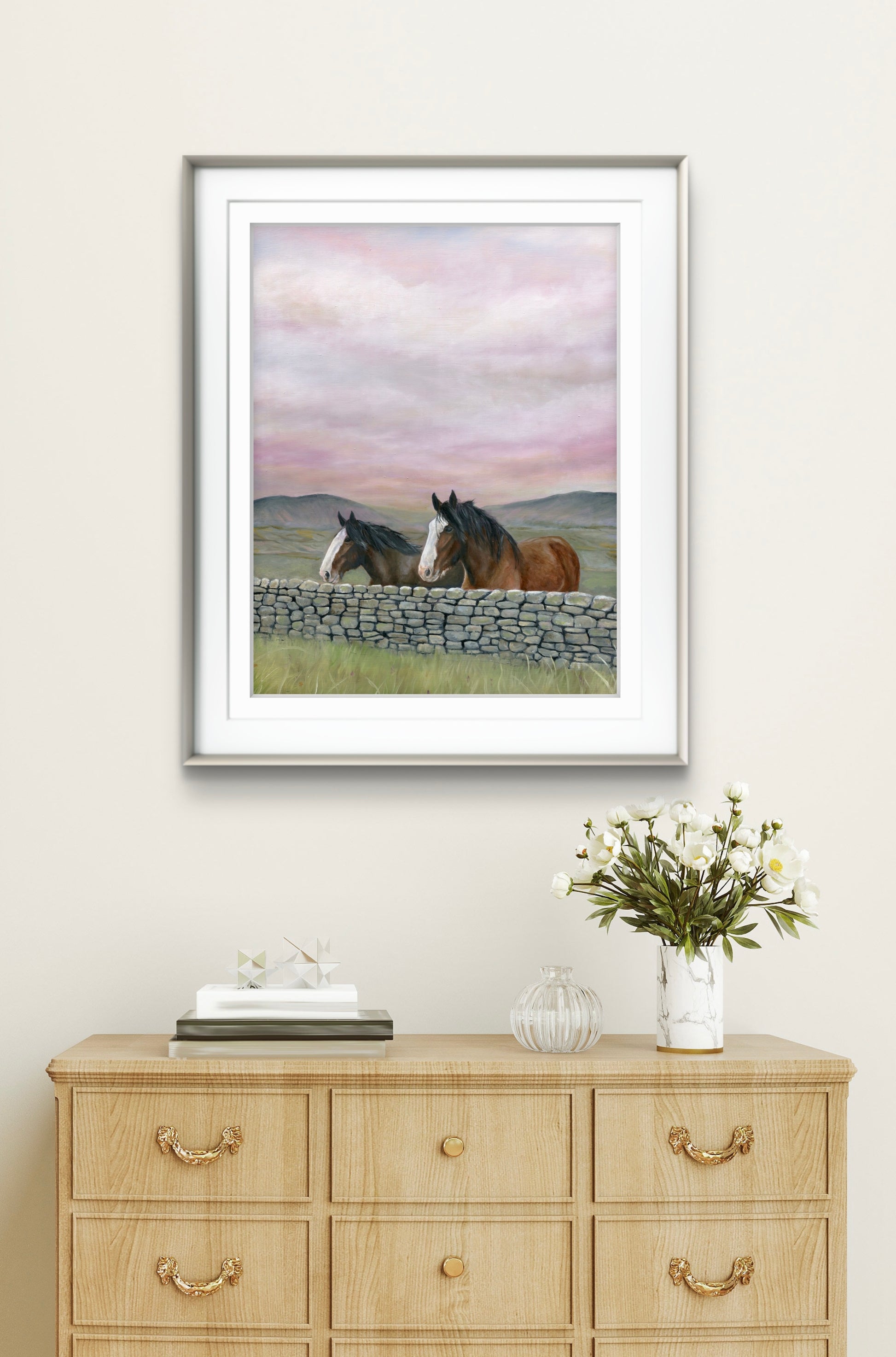 Framed artwork of two horses by a stone wall on a wall above a wooden dresser with decor items.