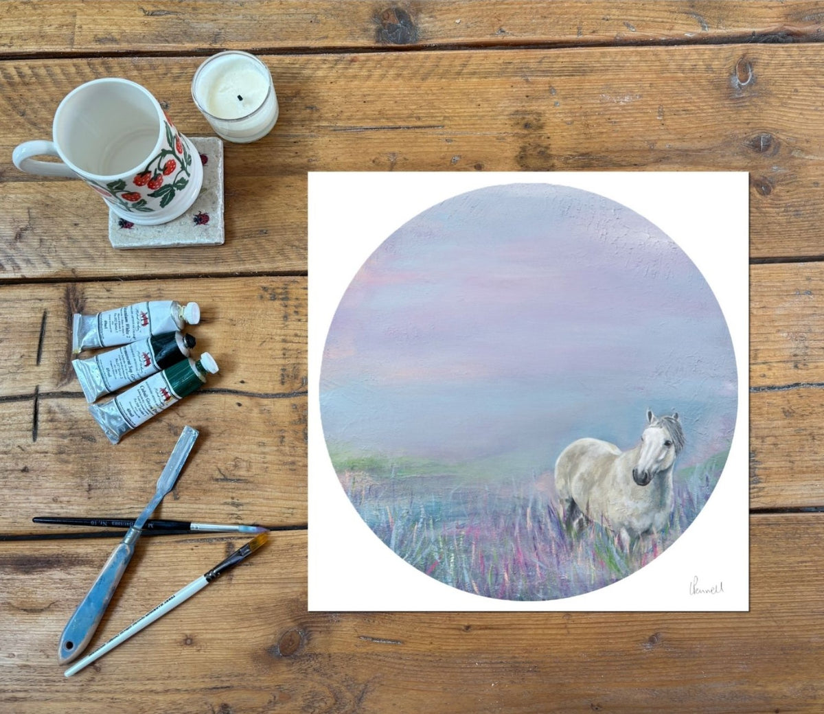 A square art print featuring a pastel-style painting of a white horse standing in a lavender and pink landscape, displayed on a rustic wooden table alongside a ceramic mug, candle, paint tubes, and paintbrushes.