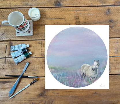 A square art print featuring a pastel-style painting of a white horse standing in a lavender and pink landscape, displayed on a rustic wooden table alongside a ceramic mug, candle, paint tubes, and paintbrushes.