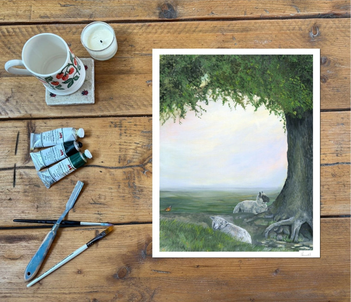 Fine art print depicting sheep in a countryside setting under an oak tree. On a wooden table with paints and brushes by the side