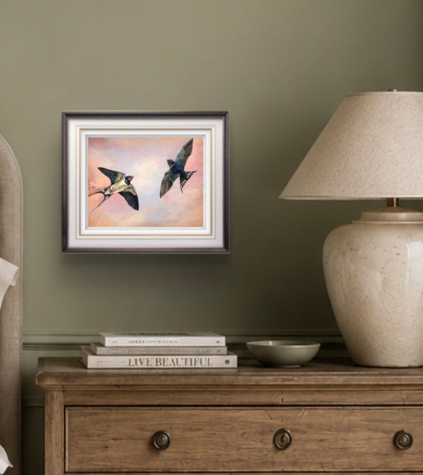Framed art print of swallows on a wall above a wooden dresser with books and a lamp.