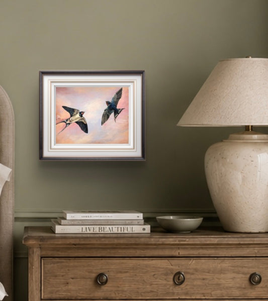 Framed art print of swallows on a wall above a wooden dresser with books and a lamp.