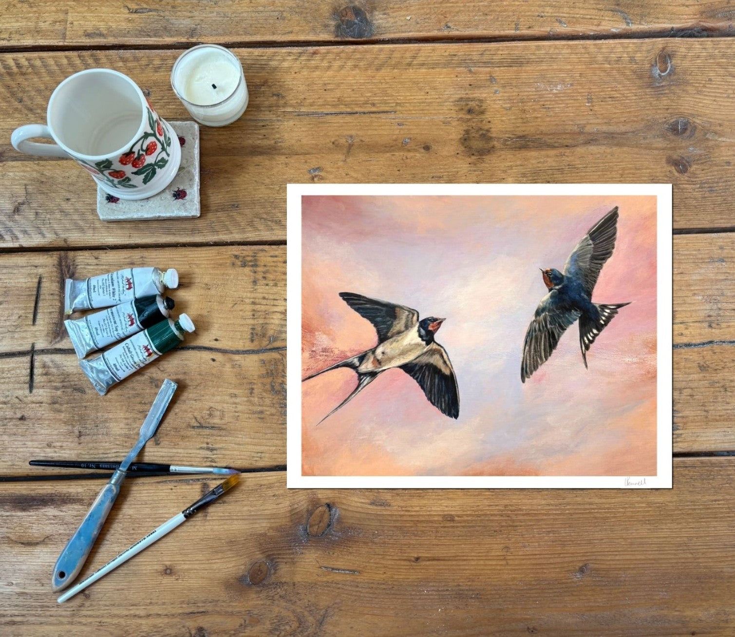 A square art print depicting two flying swallows against a soft pink and peach sky, styled in a flat-lay on a rustic wooden table alongside painting supplies including a mug, candle, paint tubes, and brushes.