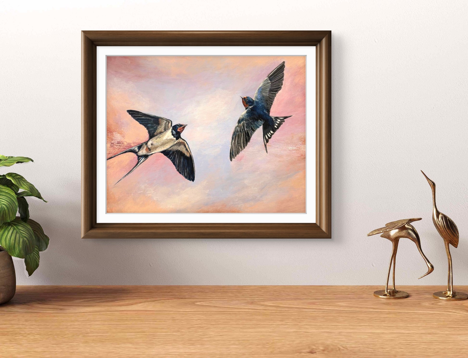 Framed artwork of two swallows flying against a pink sky, displayed on a wall above a wooden surface with decorative items.
