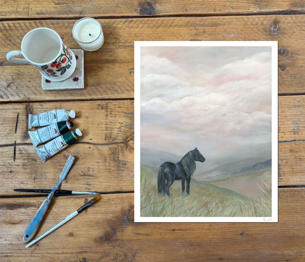 A framed art print depicting a black horse standing in a grassy field under a cloudy sky, styled on a rustic wooden table alongside painting supplies including a mug, candle, paint tubes, and brushes.