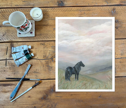 A framed art print depicting a black horse standing in a grassy field under a cloudy sky, styled on a rustic wooden table alongside painting supplies including a mug, candle, paint tubes, and brushes.