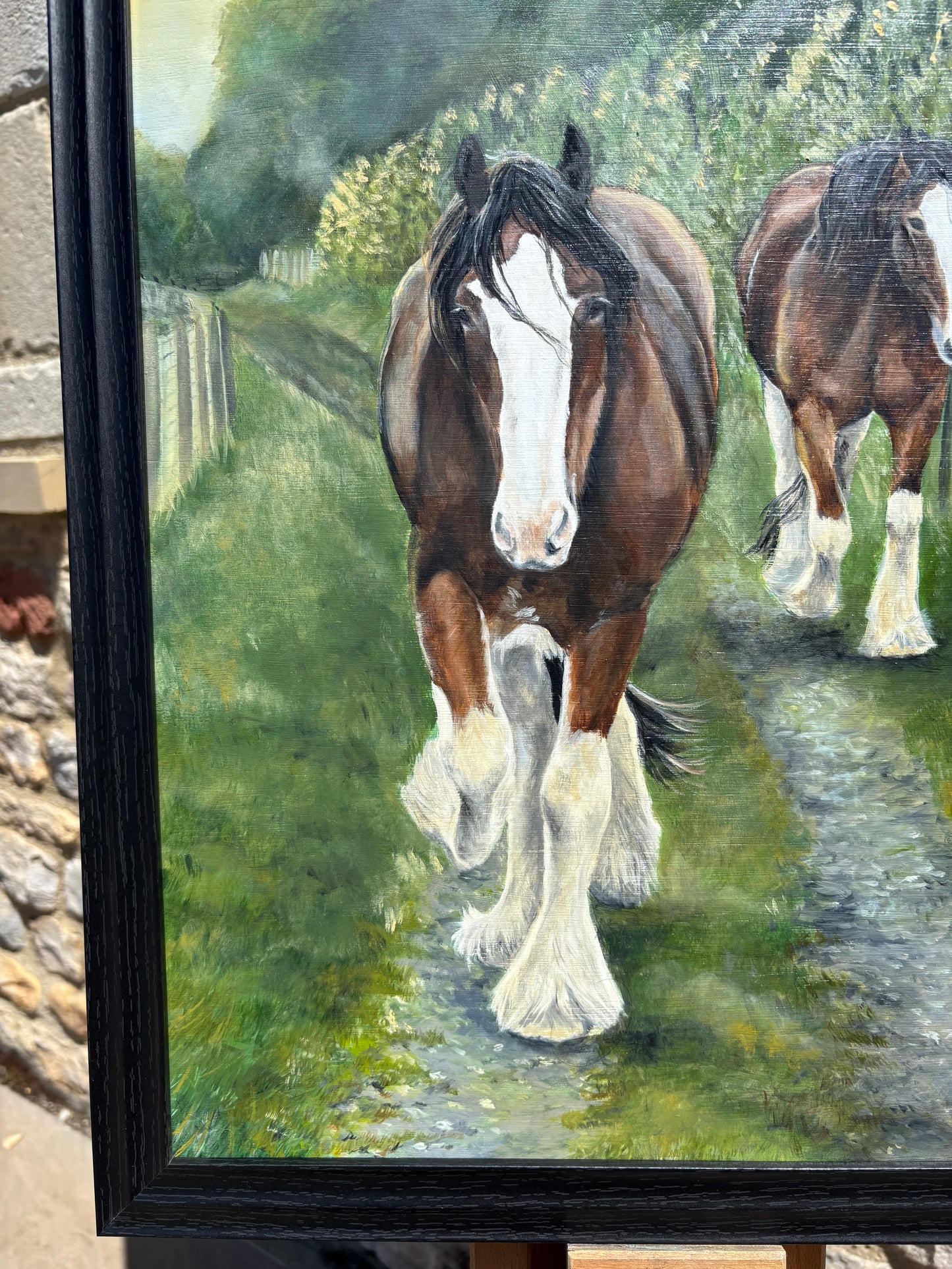 A framed painting of two brown and white draft horses walking along a grassy path, shown in a black frame on an easel.