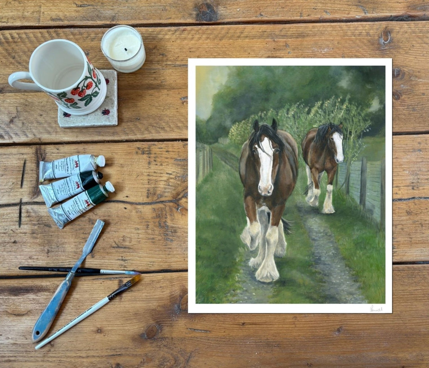 A square art print depicting two brown and white Clydesdale horses walking along a path in a green field, displayed on a rustic wooden table alongside painting supplies including a mug, candle, paint tubes, and brushes.