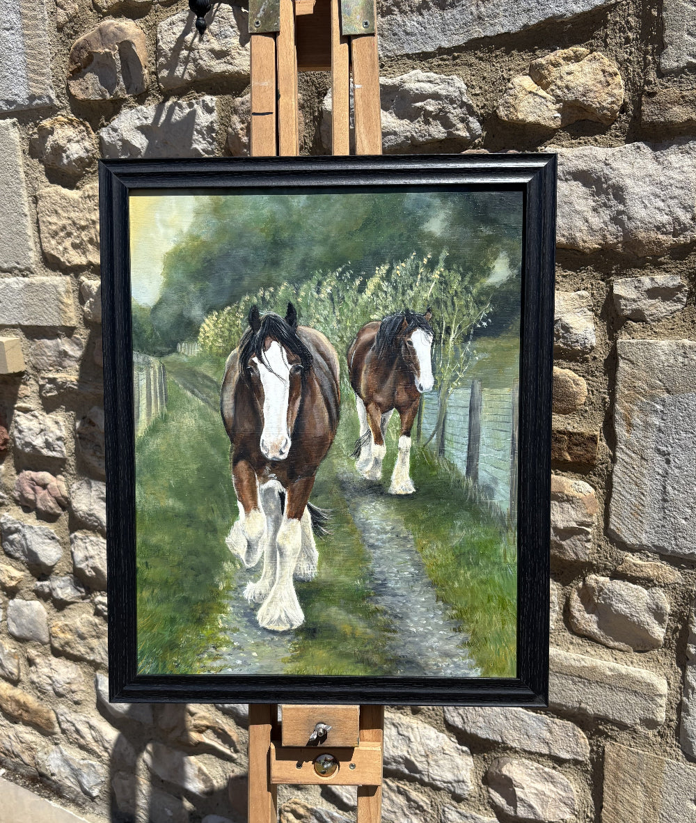 A framed painting of two brown and white horses walking along a path in a grassy field, displayed on a wooden easel against a stone wall.