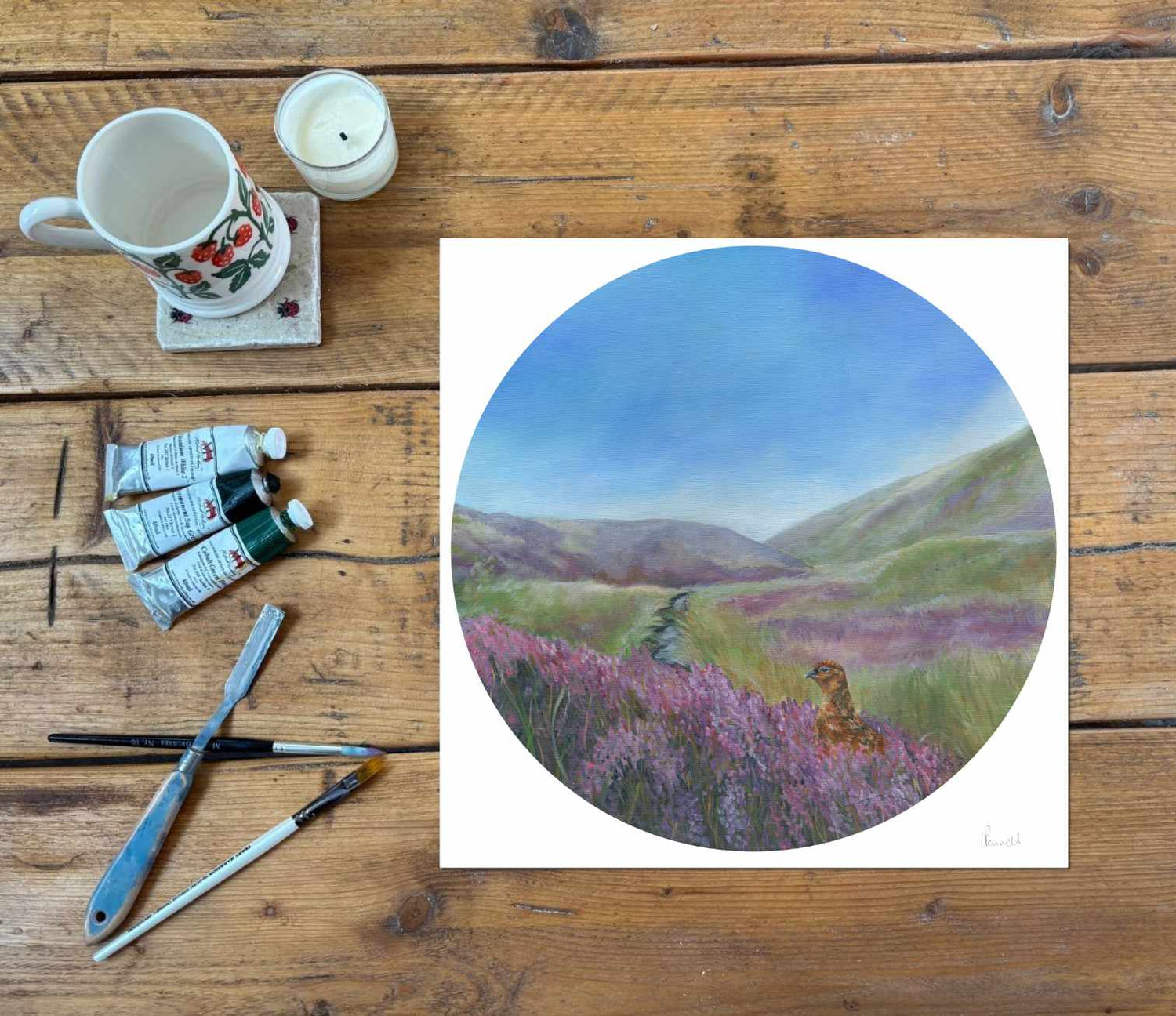 A circular pastel landscape fine art print, depicting purple heather fields and green hills under a blue sky, styled on a rustic wooden table with paint tubes, brushes, a mug, and a candle.
