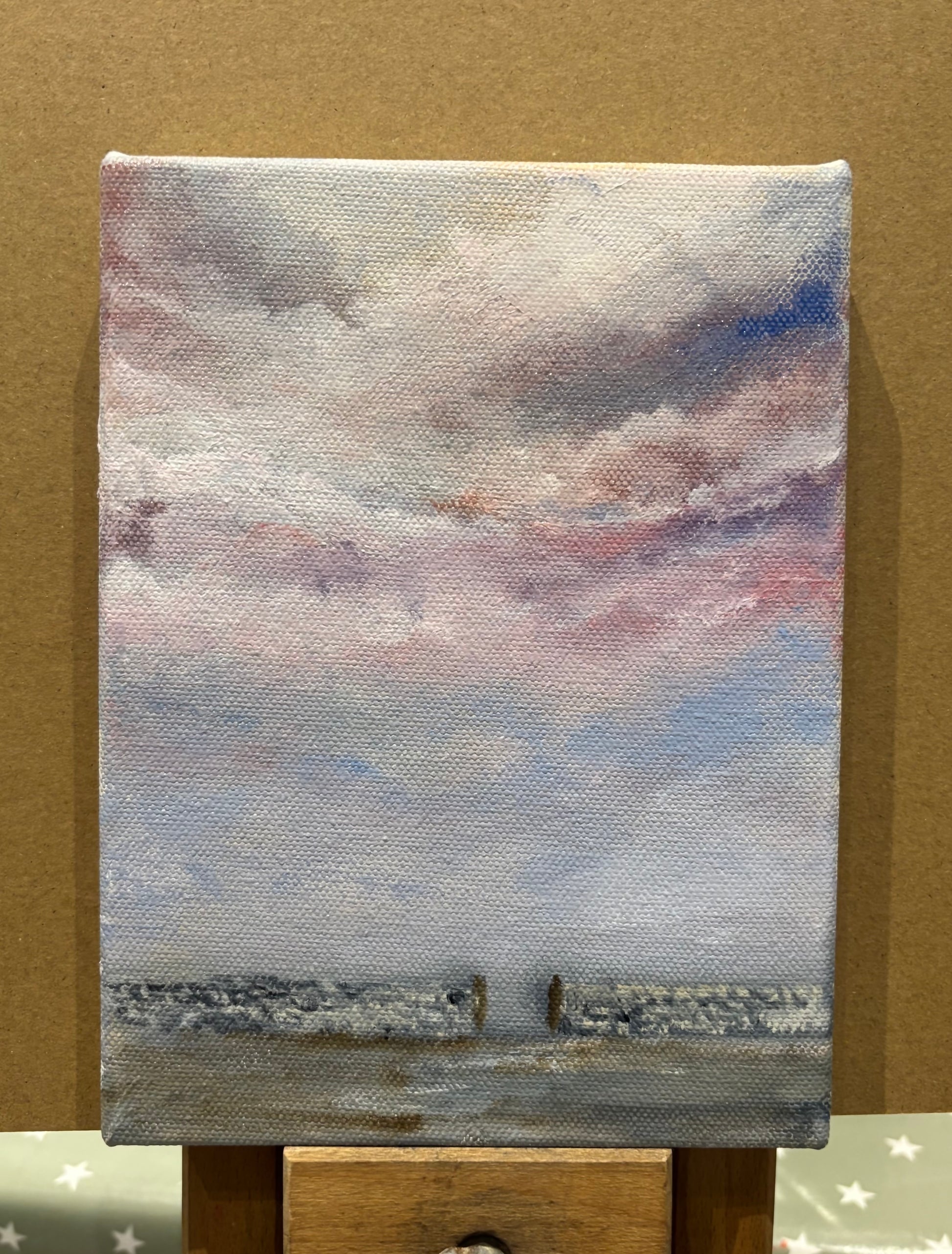 Small painting of a landscape with a cloudy sky against a brown background perched on an easel