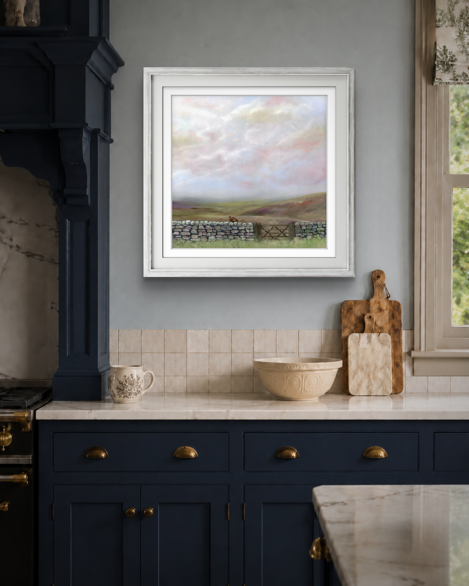Framed landscape painting above a kitchen counter with blue cabinets and a window in the background.