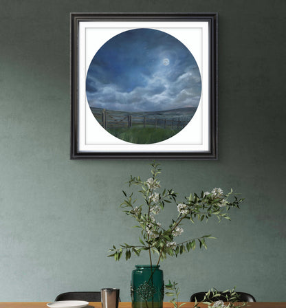Framed artwork of a landscape with a fence and sky, displayed on a wall above a table with a vase and branches.