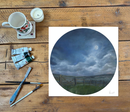 A circular art print depicting a moonlit night sky over a moorland grassy field and fence, displayed on a rustic wooden table alongside painting supplies including a floral ceramic mug, candle, paint tubes, and brushes.
