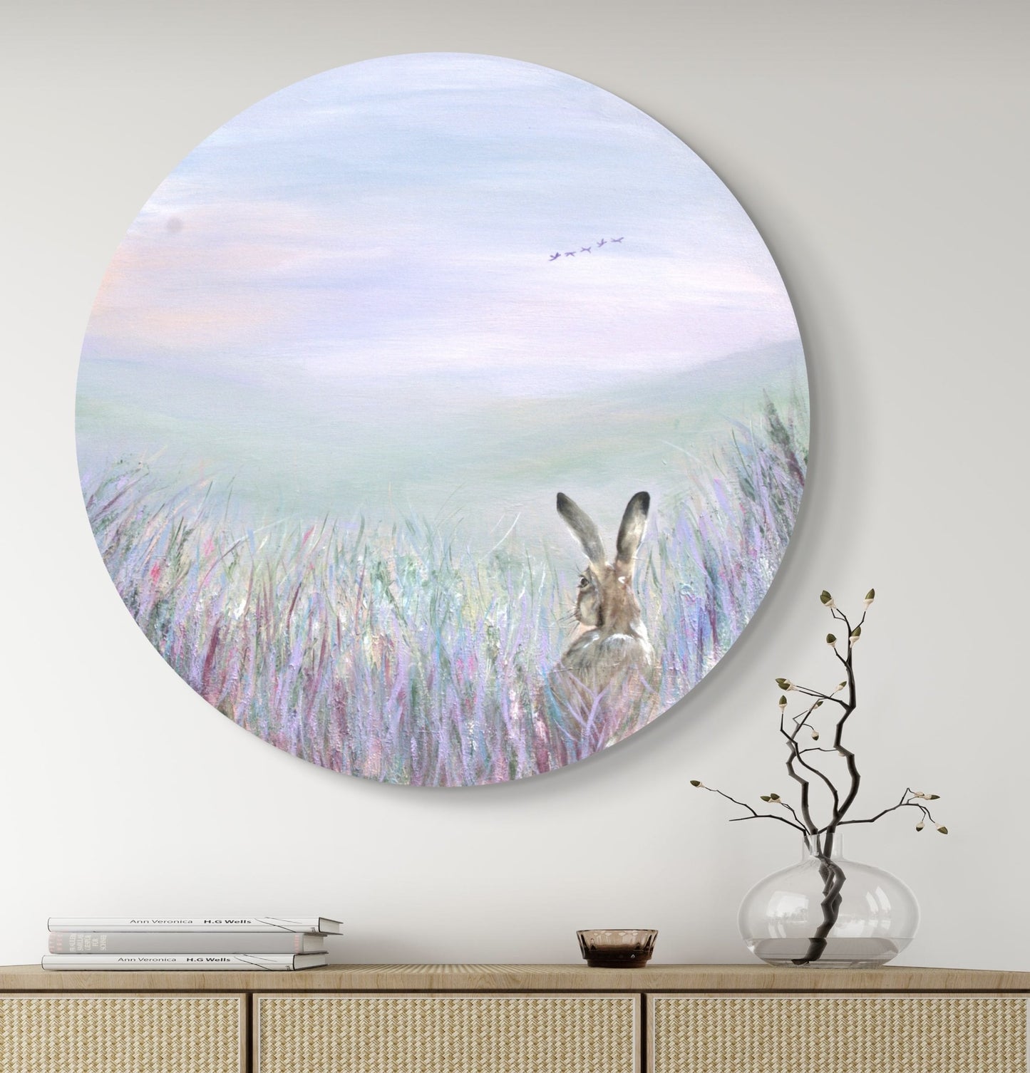 An acrylic painting depicting a hare peeping out through a wildflower border, with a vast sweeping landscape in the background. The artwork is  displayed on a wall above a furniture settee, with a vase of dried flowers to the side.