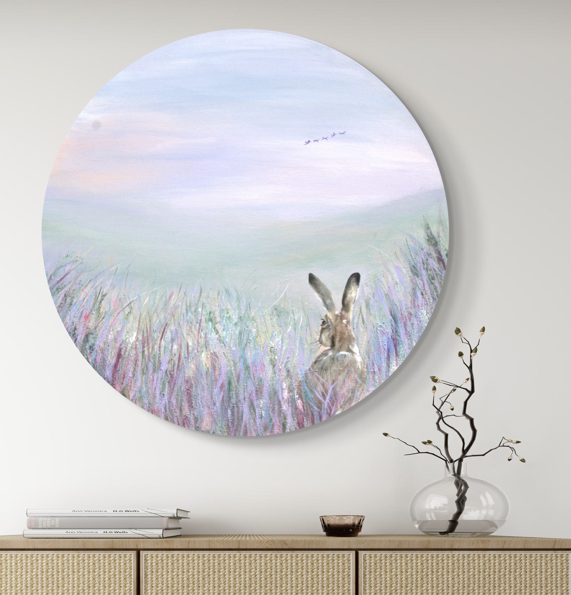 An acrylic painting depicting a hare peeping out through a wildflower border, with a vast sweeping landscape in the background. The artwork is  displayed on a wall above a furniture settee, with a vase of dried flowers to the side.