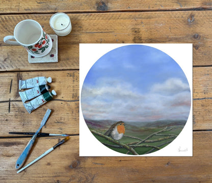 A circular art print featuring a robin bird perched on branches under a blue and white sky, displayed on a rustic wooden table alongside painting supplies including a mug, candle, paint tubes, and brushes.