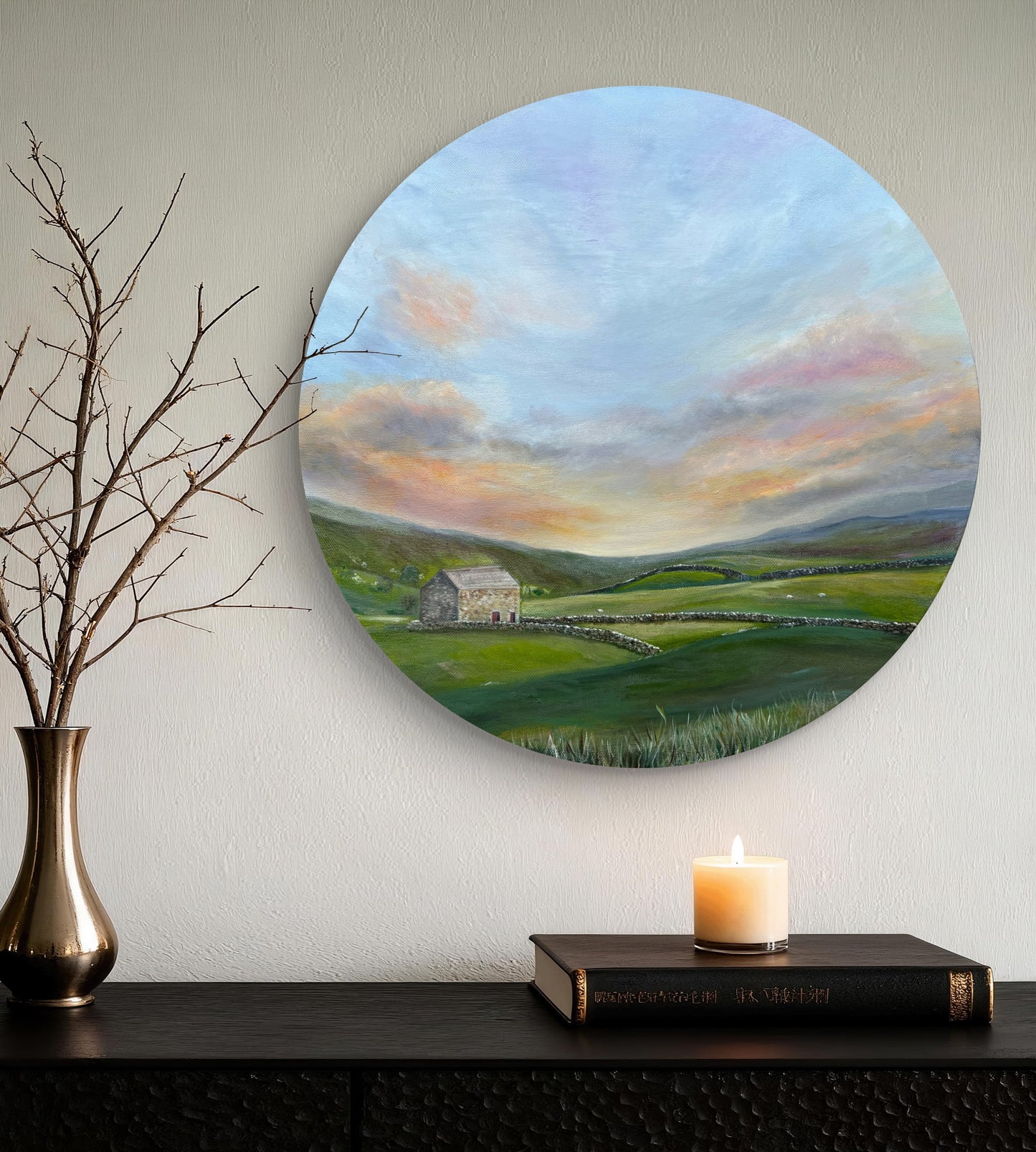 Round painting of a landscape with a house and sunset, displayed on a wall above a shelf with a candle and books.
