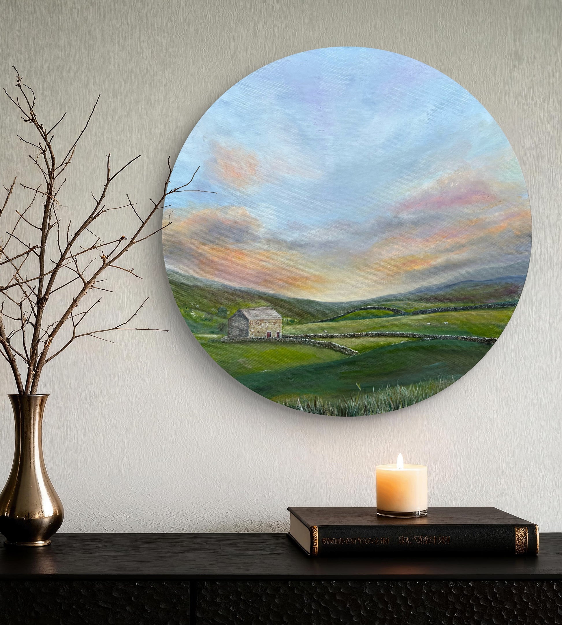 Round painting of a landscape with a house and sunset, displayed on a wall above a shelf with a candle and books.