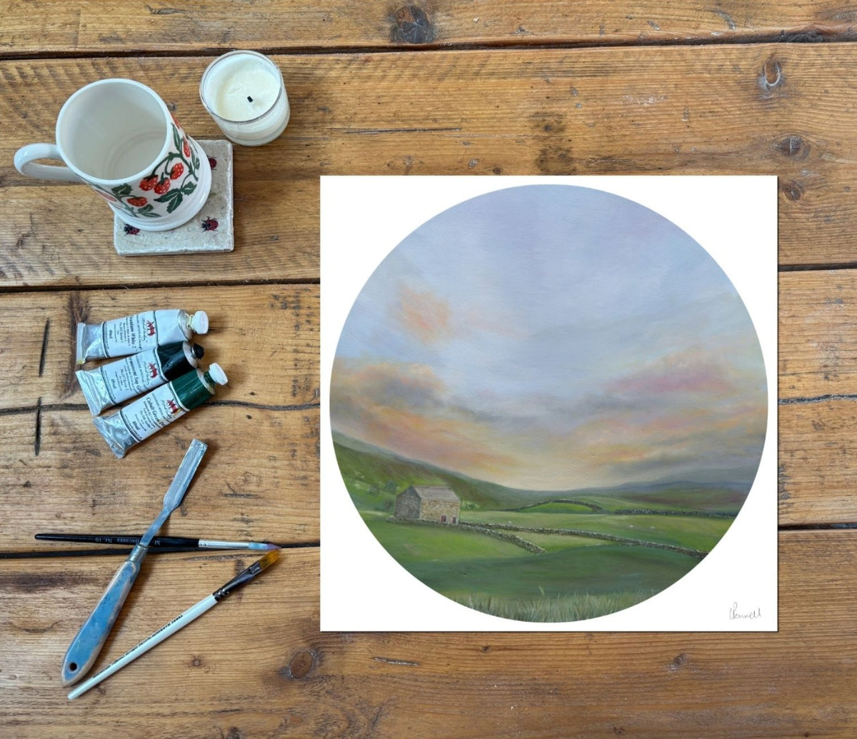 A circular print showing a Yorkshire countryside landscape with moorland and a drystone walled barn. On a rustic wood table with painting supplies next to it 