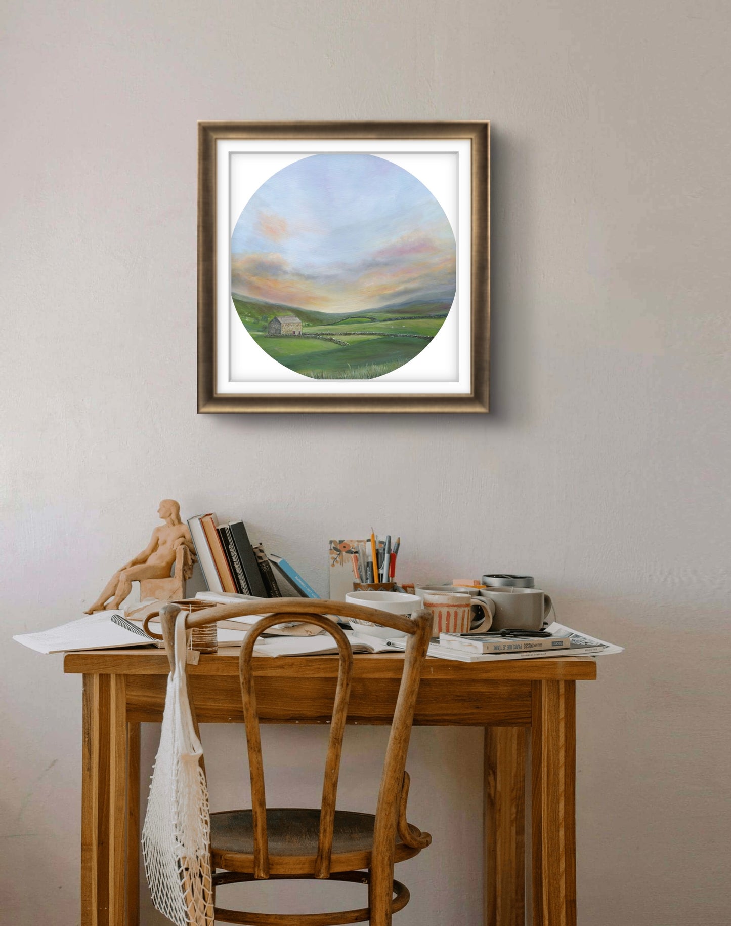 Framed circular painting of a landscape above a wooden desk with stationery items.