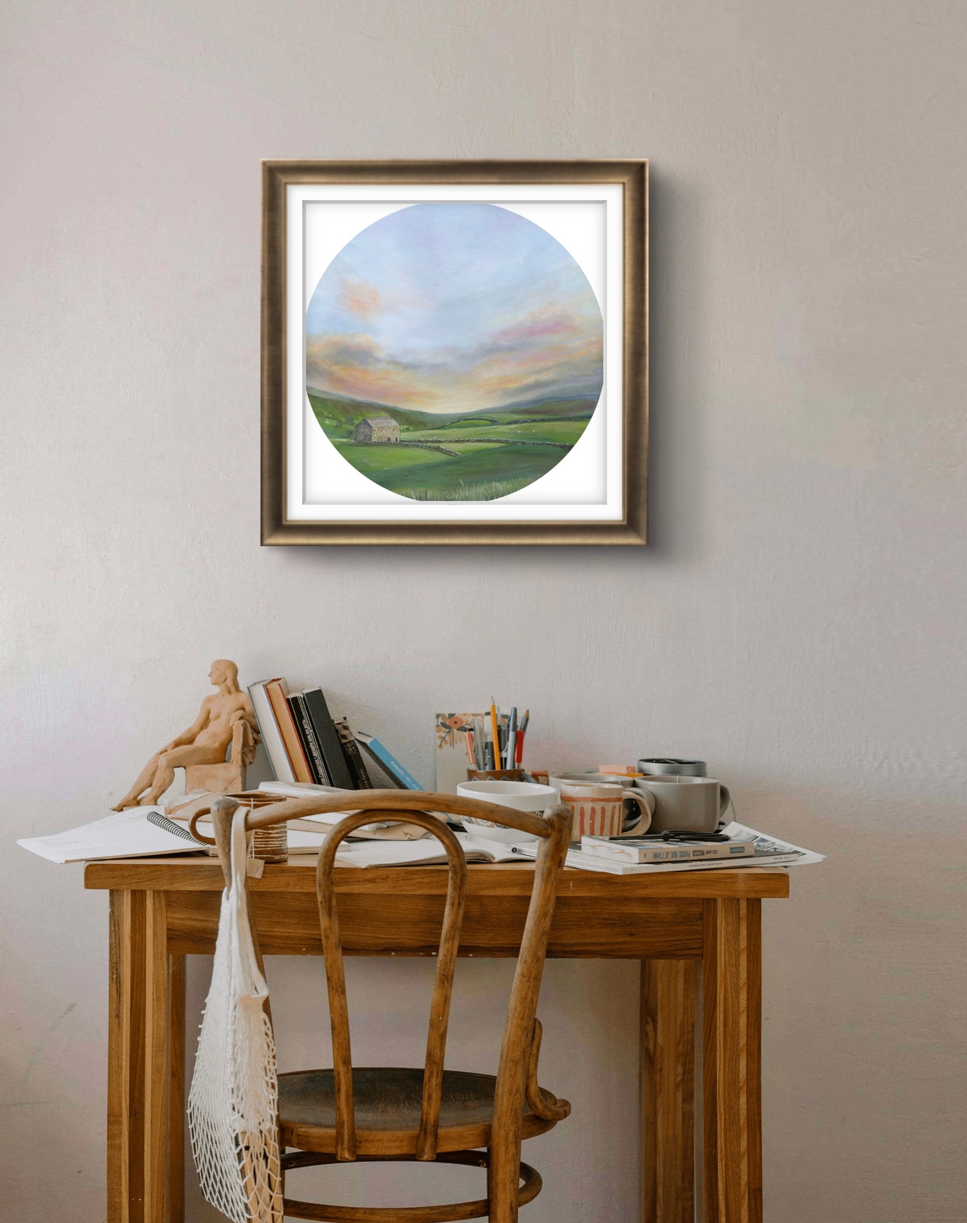 Framed circular painting of a landscape above a wooden desk with stationery items.