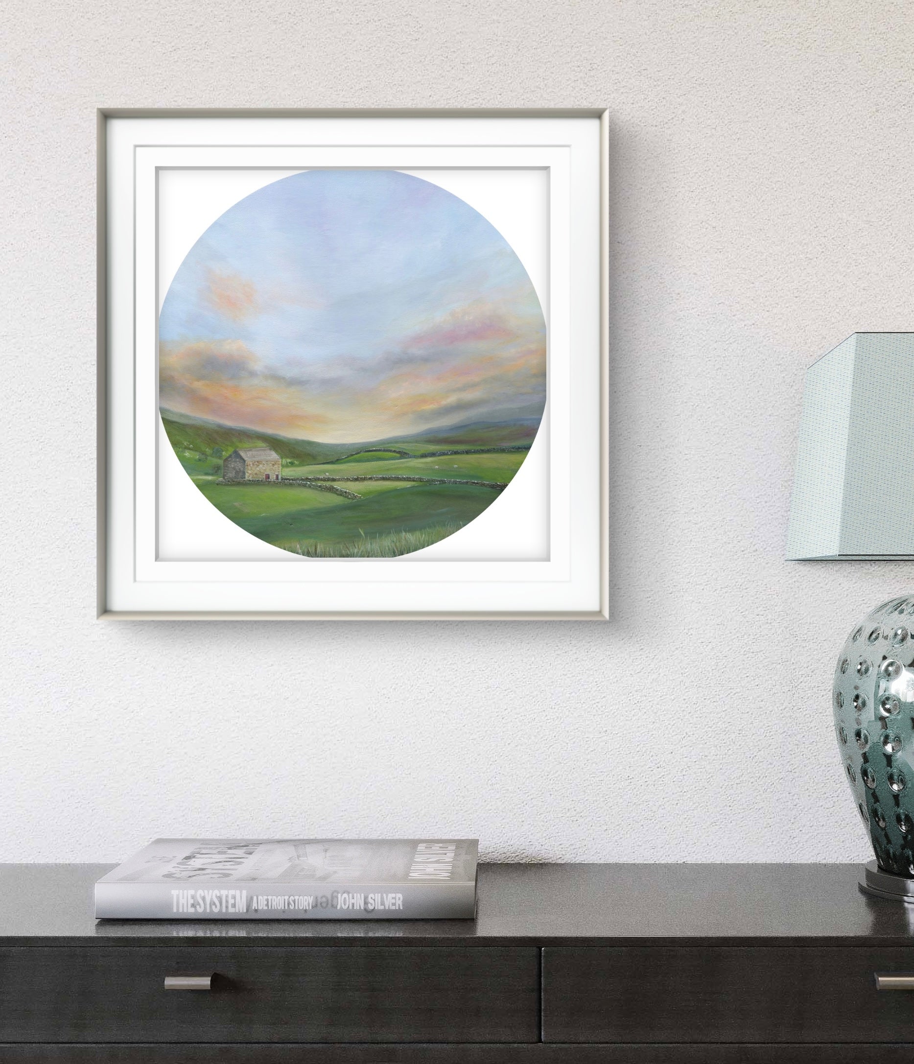 Framed artwork of a landscape with a house on a wall above a table.