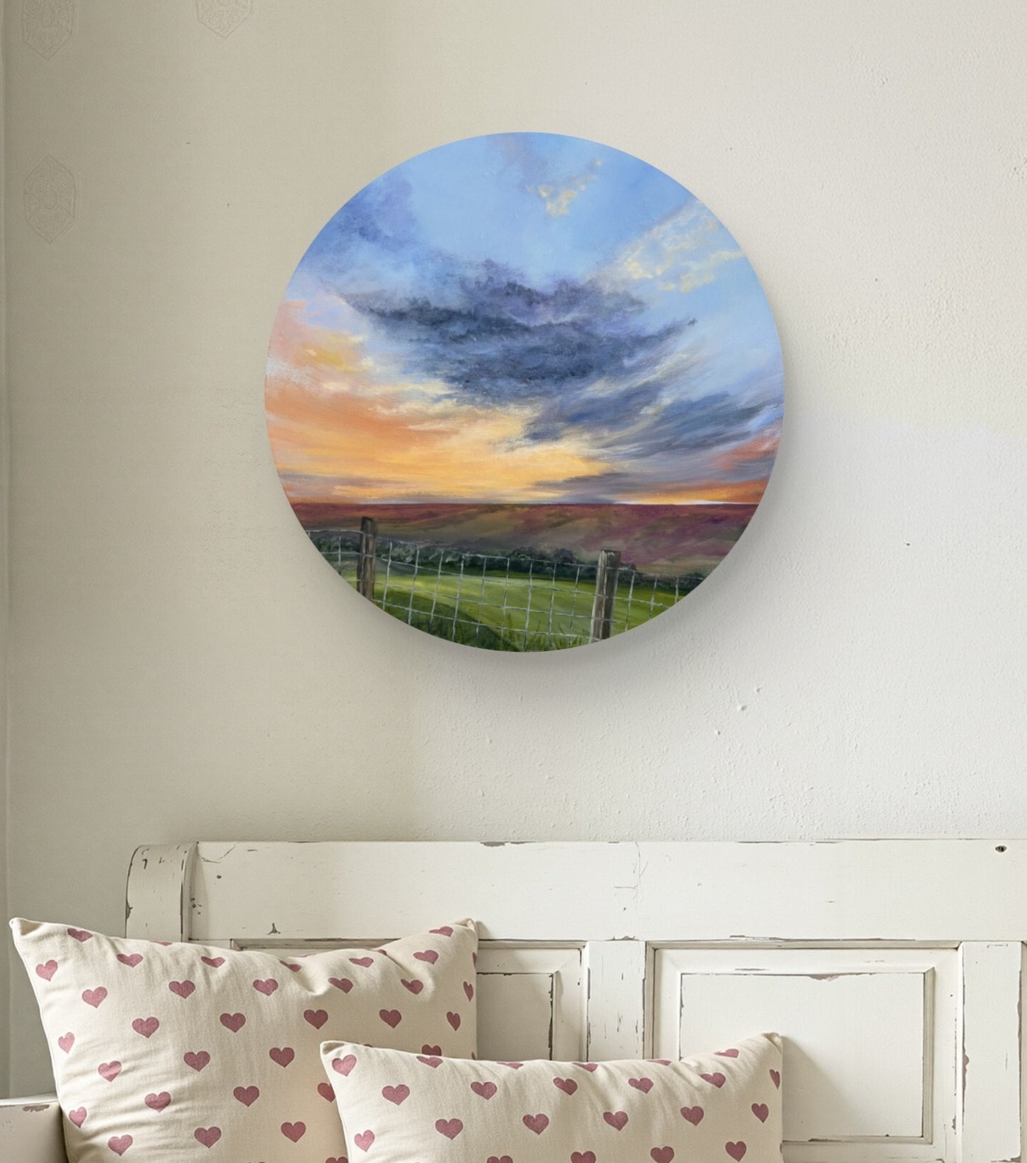 Round wall art depicting a sunset over a field, hanging above a bed with heart-patterned pillows.
