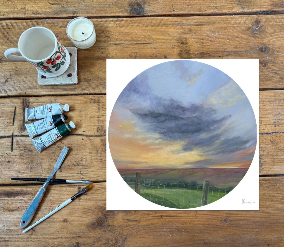 A yorkshire countryside scene with a big cloudy sky, in a circular print, on a rustic wood table with painting supplies by the side
