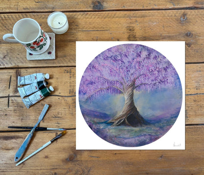 A purple tree depicted in a circle on a fine art print, shown on a rustic wood table with painting supplies, a mug and a candle next to it 