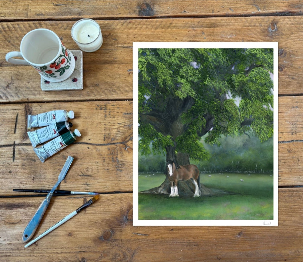 A fine art print of a clydesdale under an oak tree in a field, shown on a rustic wood table with paints, brushes, a mug and a candle by the side
