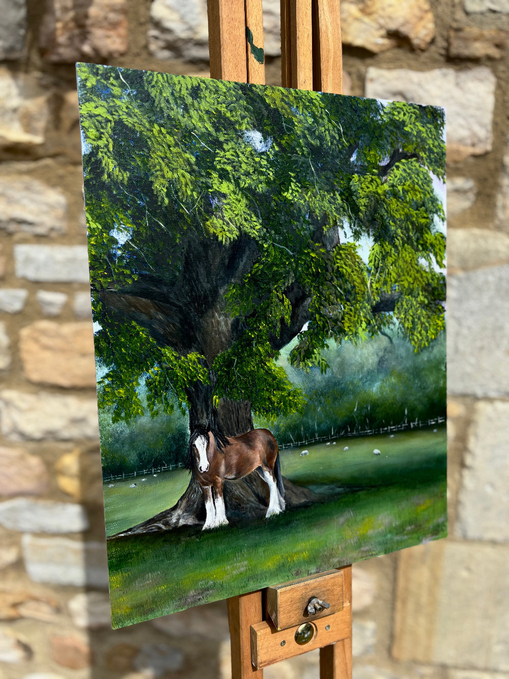 Painting of a horse under a tree on an easel against a stone wall.