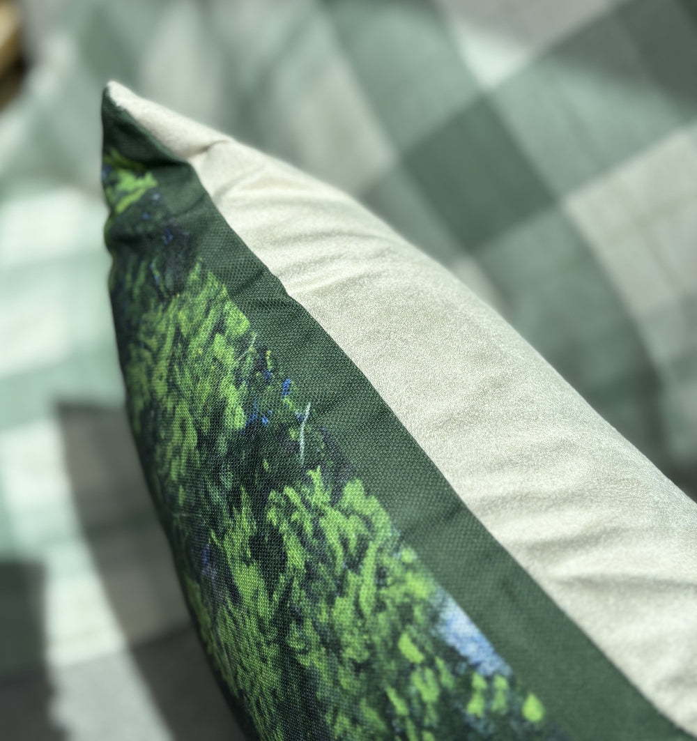 A close-up of a decorative throw pillow featuring a green forest pattern on one side and a light beige fabric border on the other, resting on a green and white checkered sofa.