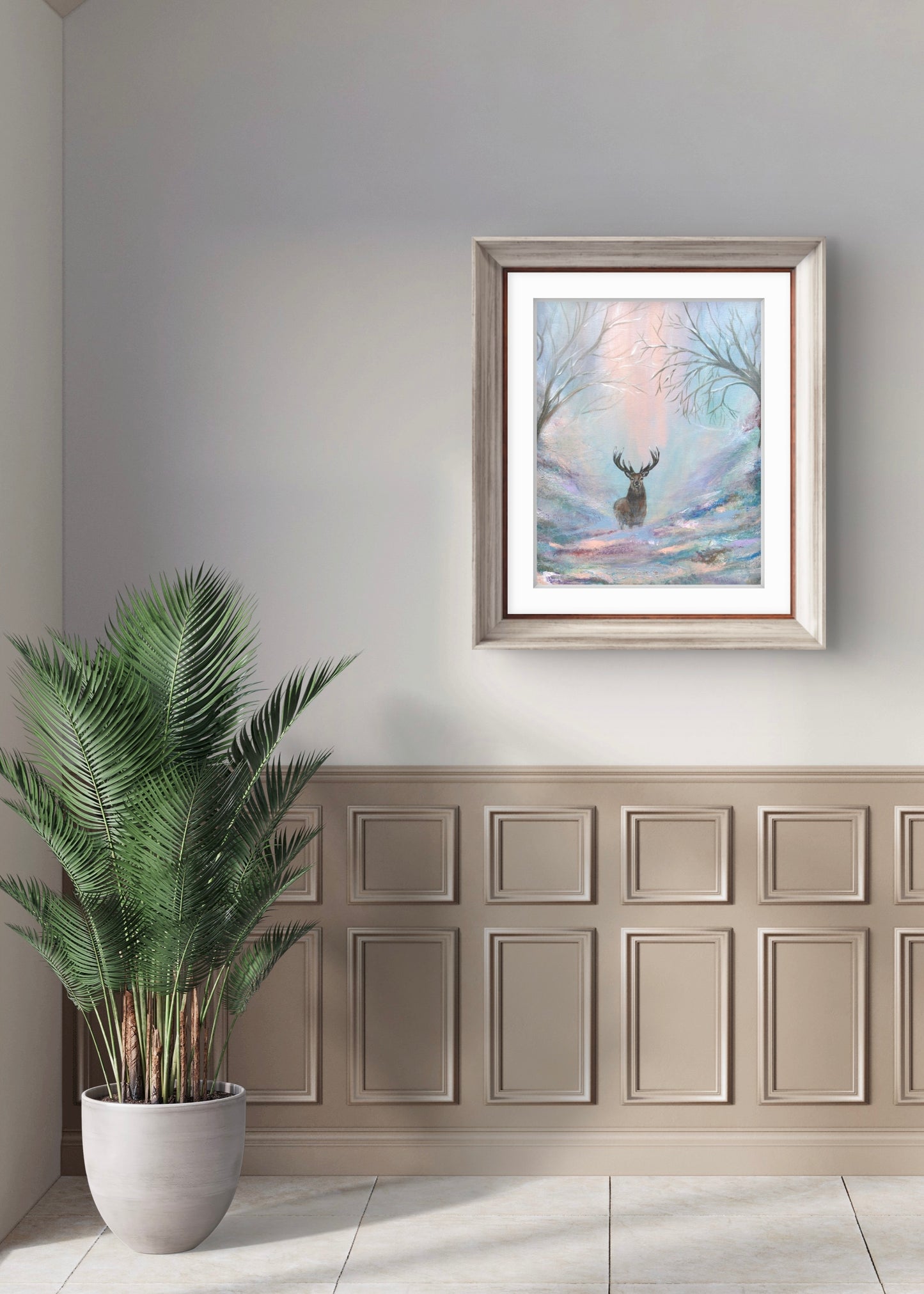 Framed artwork of a deer in a snowy landscape on a wall above a wooden paneling with a plant in the foreground.