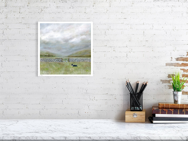 Framed moorland landscape print of a field with a sheepdog on a white brick wall above a desk.