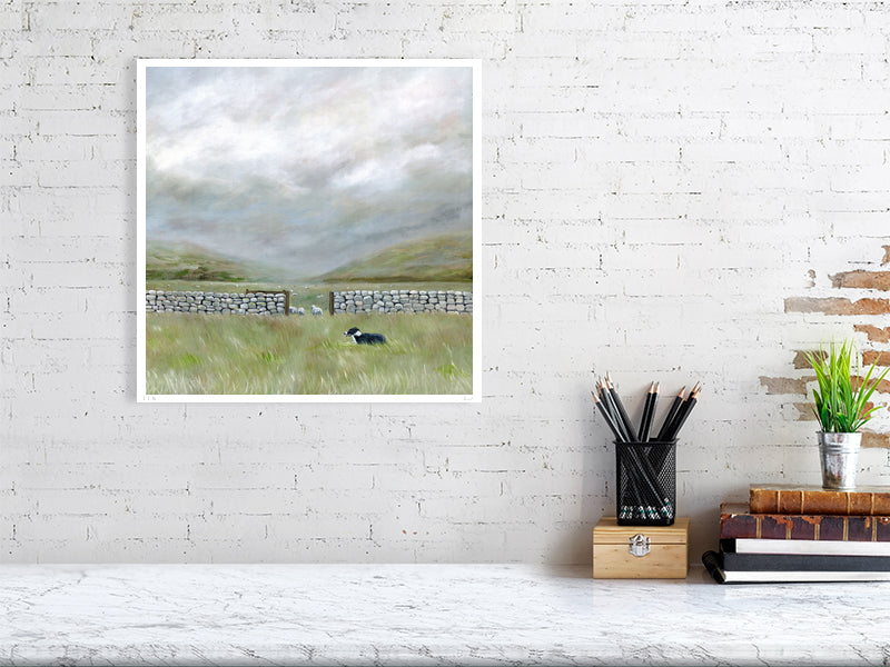 Framed landscape painting of a moorland field with a sheepdog on a white brick wall above a desk.