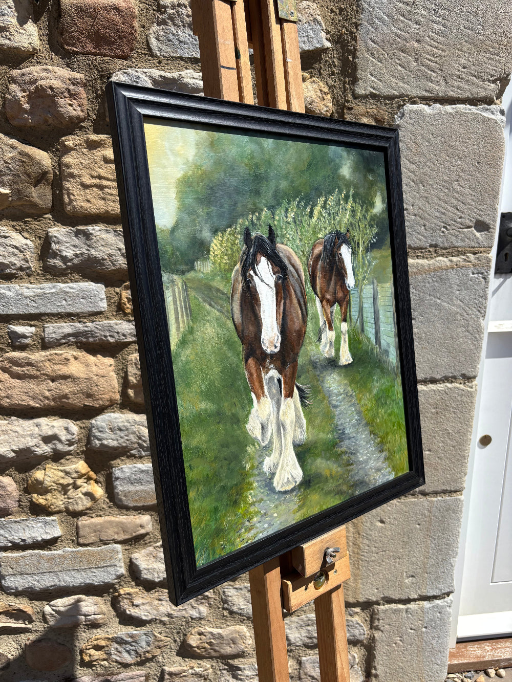 A framed painting of two horses walking along a path in a green field, displayed on a wooden easel against a stone wall.