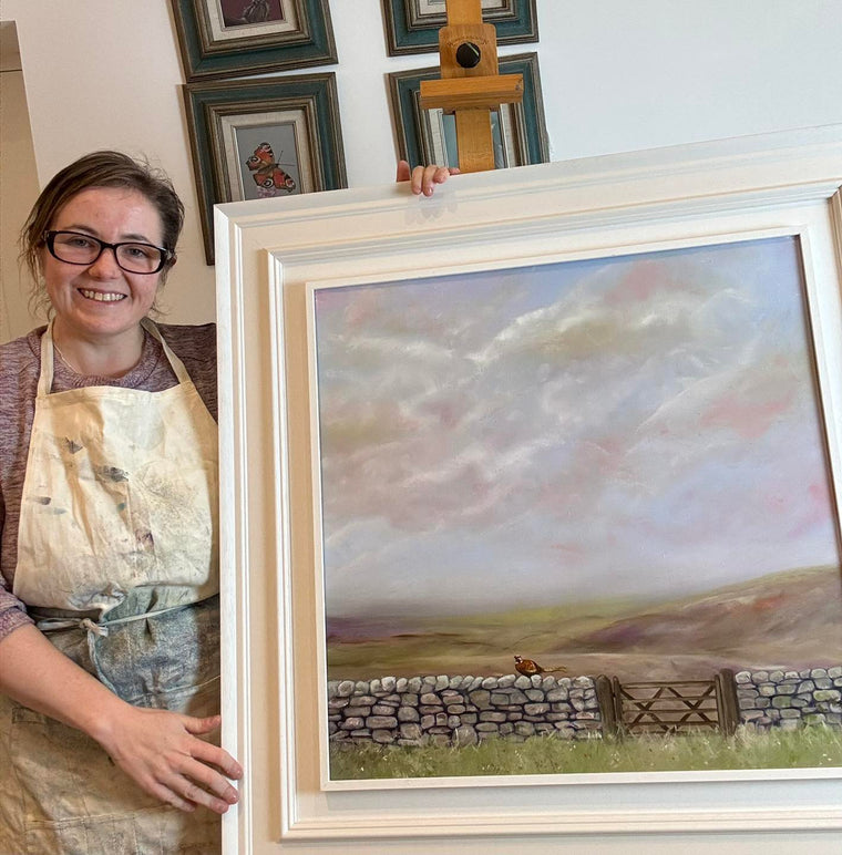 Woman holding a framed landscape painting with a stone wall and field.
